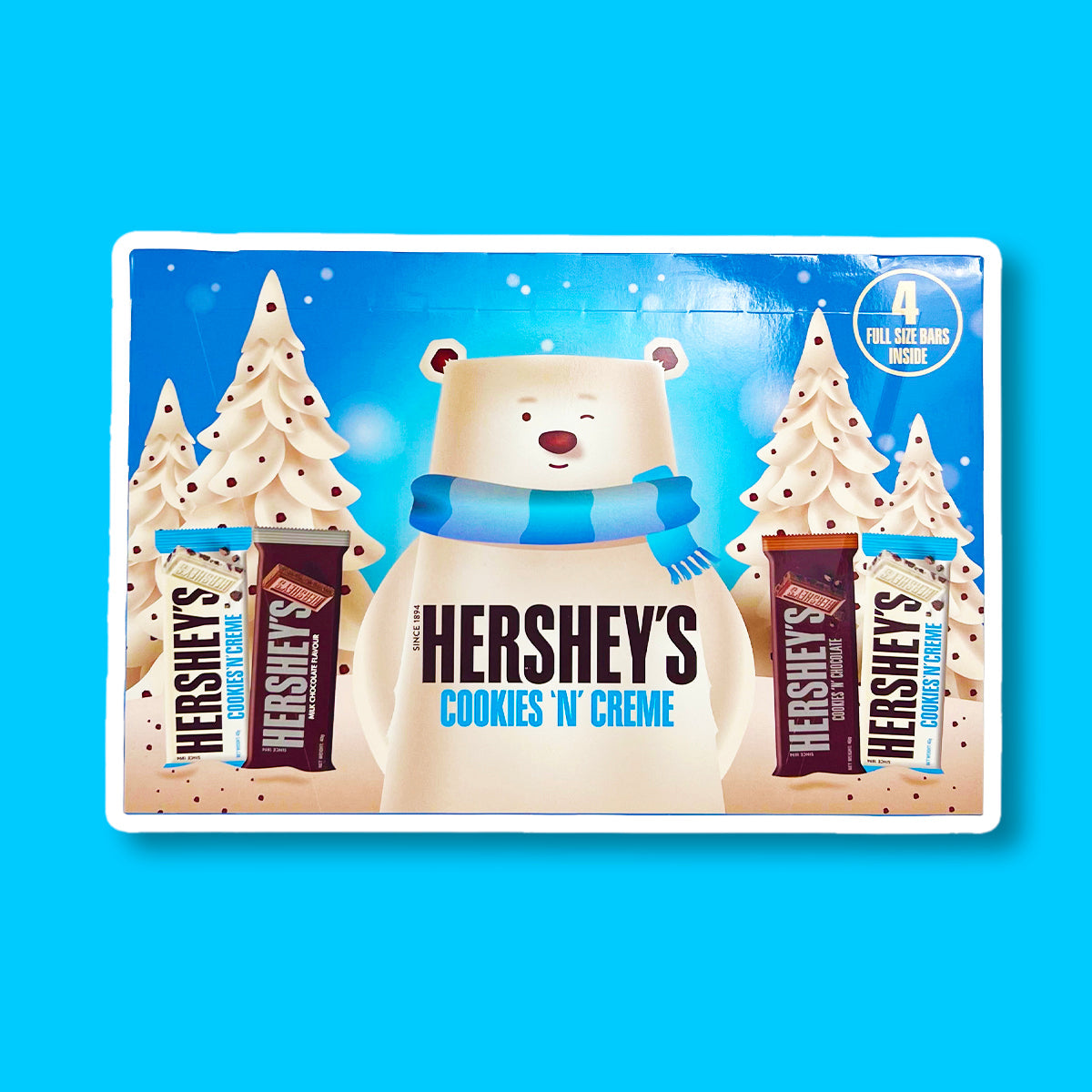 Un carton bleu sur fond bleu avec un immense ours blanc qui fait un clin d’oeil et entouré de 2 sapins blanc. De part et d’autre de l’ours, 4 barres chocolatées