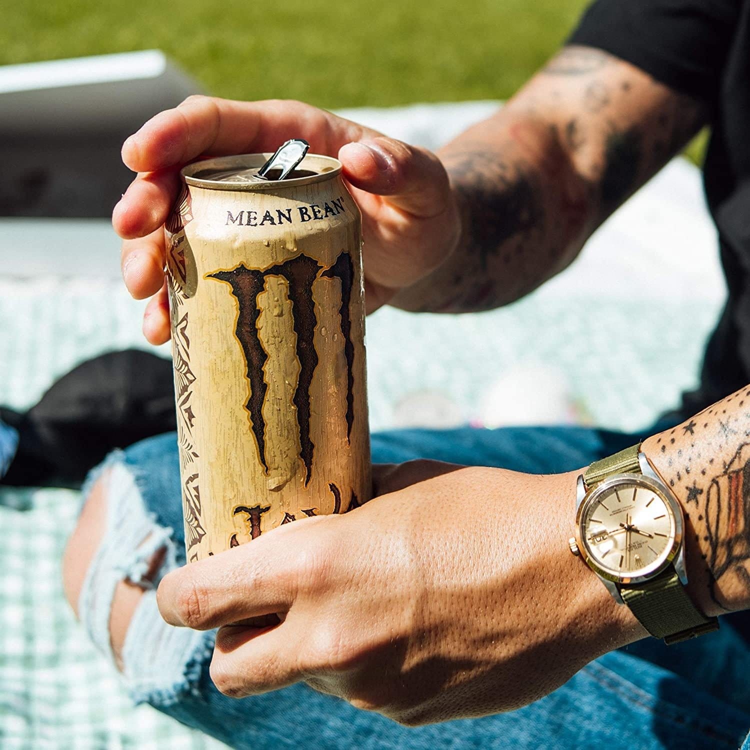 Une grande canette beige avec au centre un grand M marron dans une main avec des bras tatoués et derrière de l’herbe