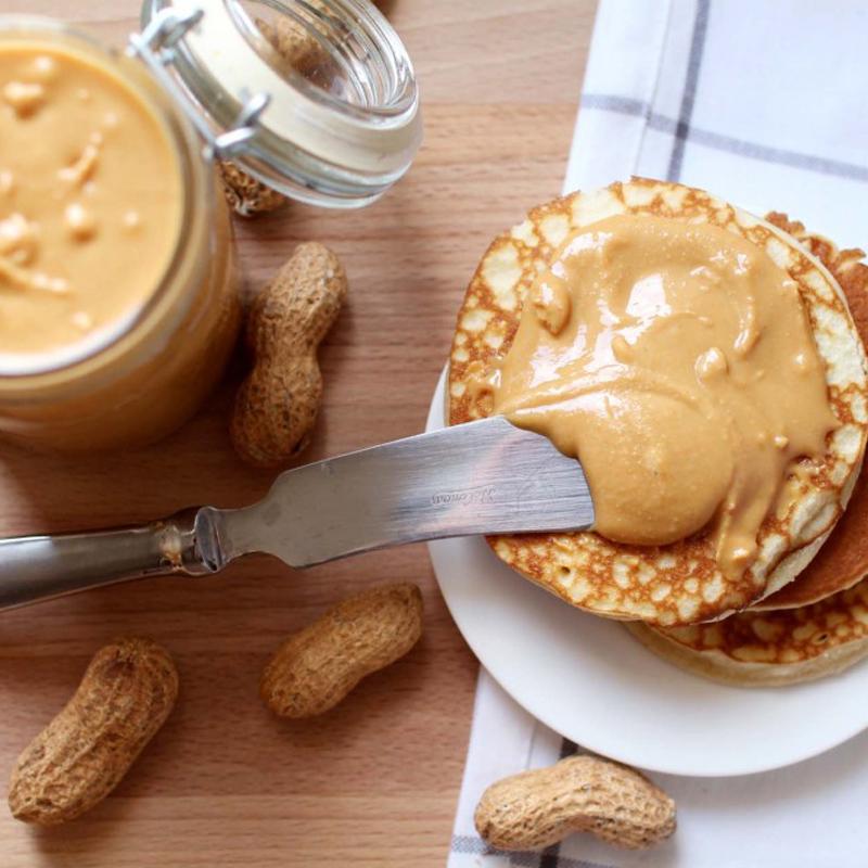 Une assiette blanche de pancakes avec une crème brune et sur la coté un pot en verre remplie de cette crème, le tout sur une table en bois avec des arachides