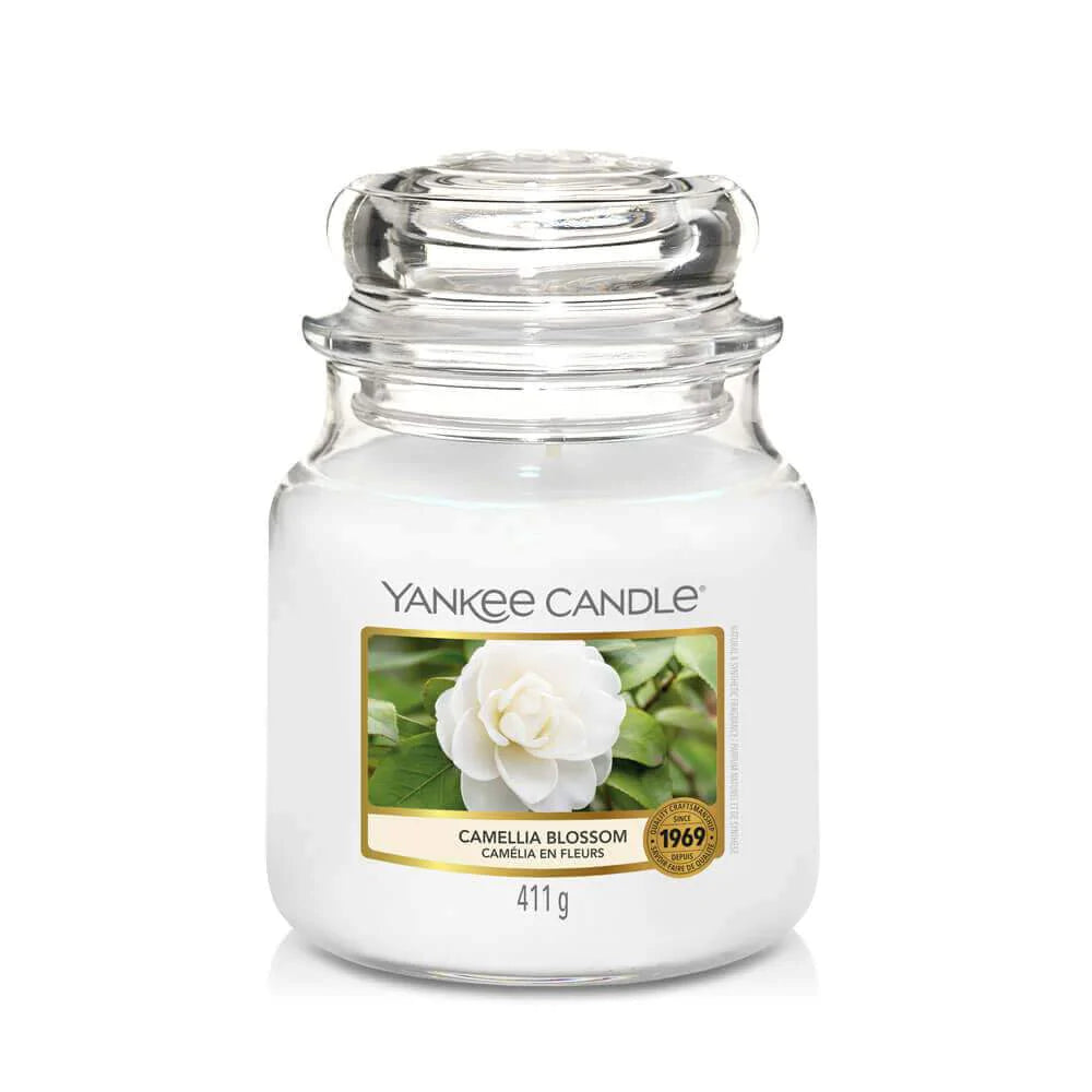 Un emballage sur fond blanc avec une bougie blanche dans un bocal en verre, un capuchon en verre et une étiquette avec une fleur blanche et des feuilles vertes à l’arrière