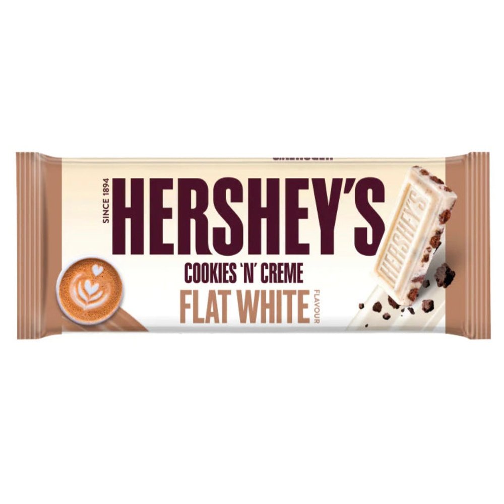 Un emballage beige avec des extrémités bruns sur fond blanc, à gauche une tasse blanche remplie de café et à droite un carré de chocolat blanc avec des morceaux de biscuits noirs