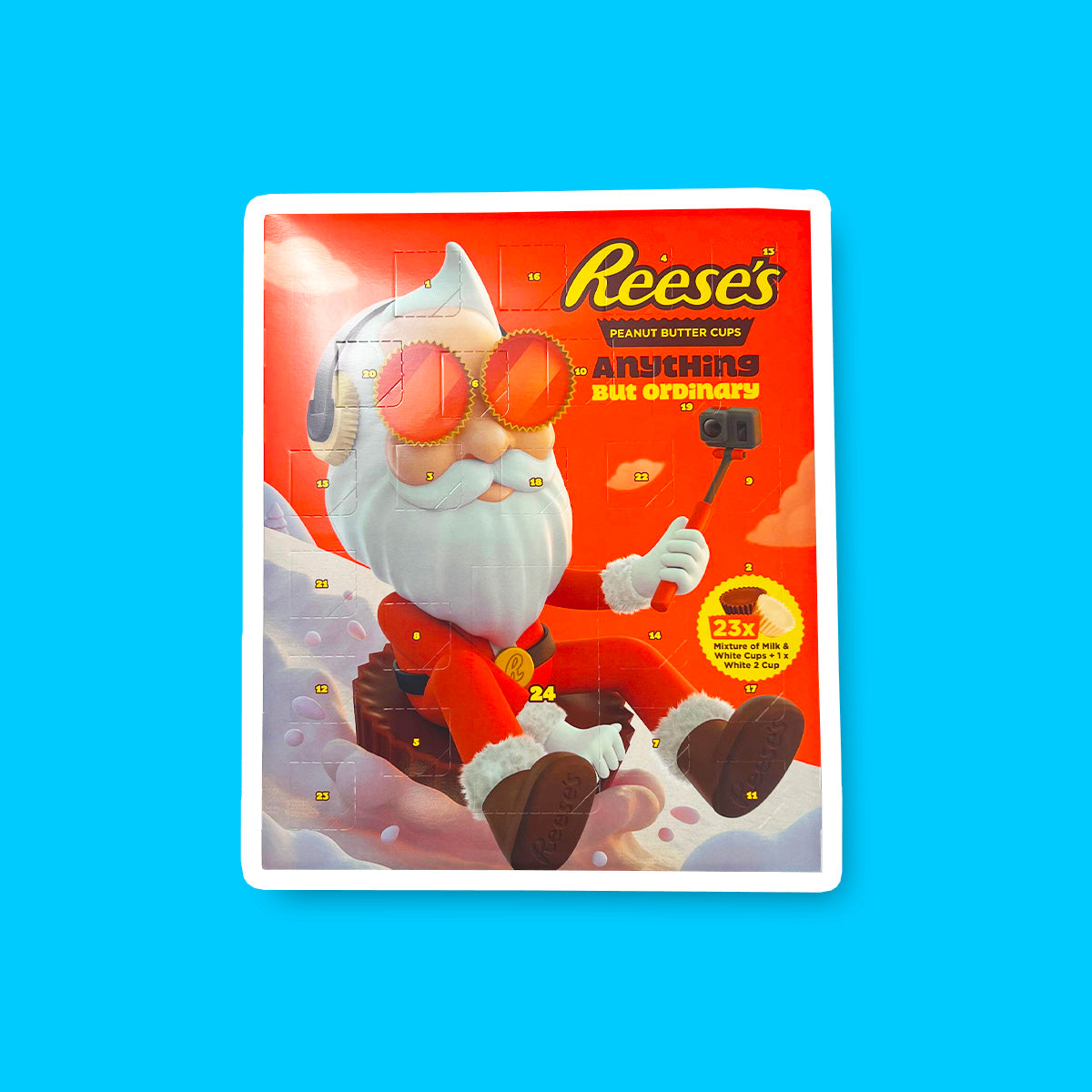 Un carton orange sur fond bleu avec un père Noël stylé qui se filme entrain de descendre une montage enneigée assis sur un chocolat Reese’s géant