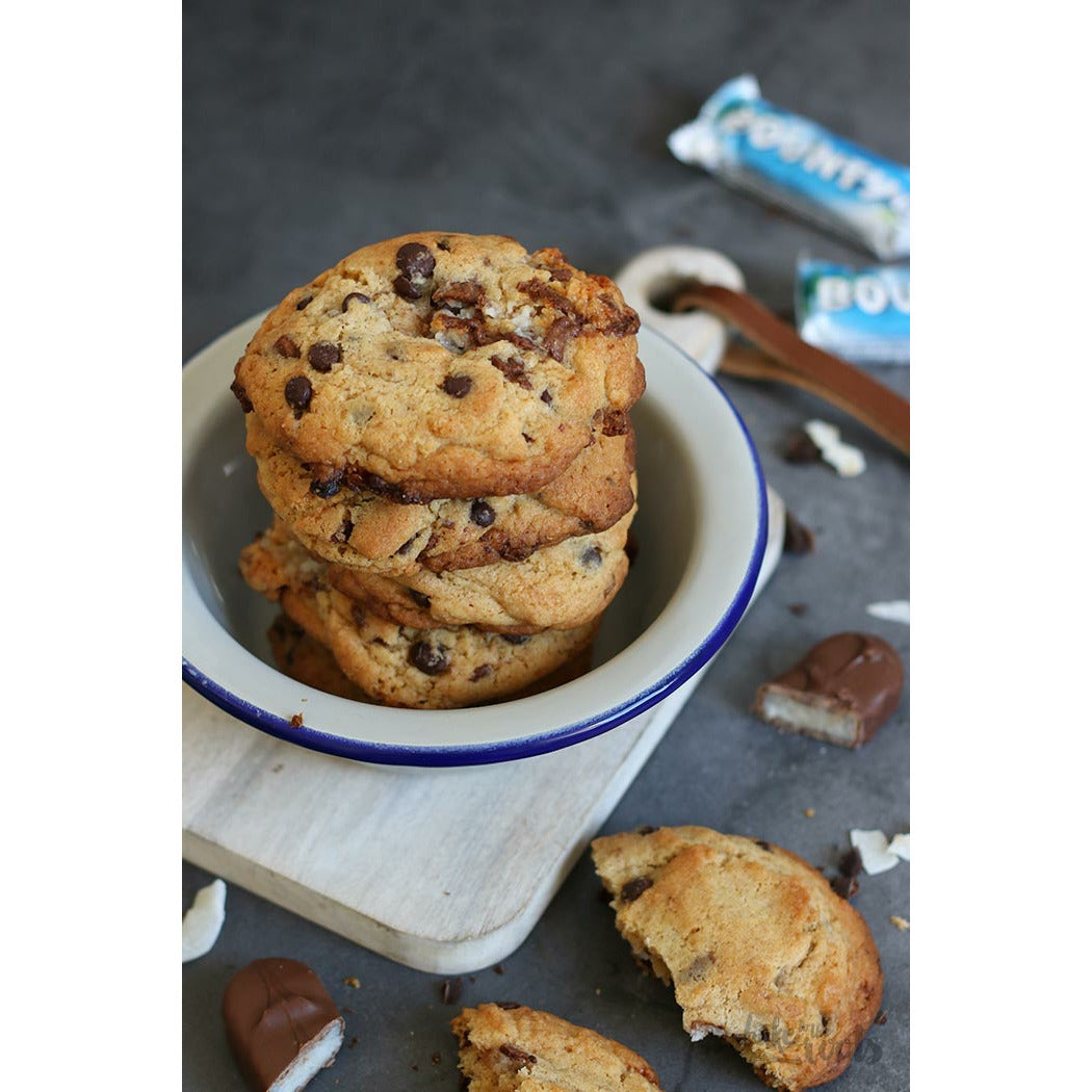 Des cookies dans une assiette blanche aux rebords bleus, sur une planche et sur une table grise. Autour il y a des bounty