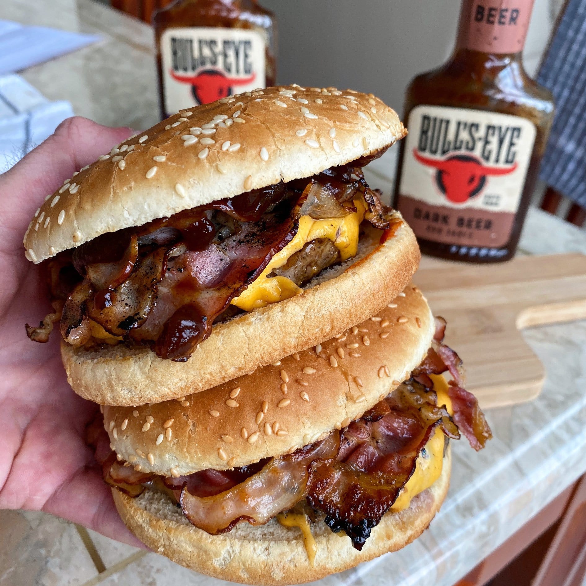 Une main qui tient 2 hambugers avec du fromages et des tranches de bacon et à l'arrière 2 bouteilles de sauve sur une table