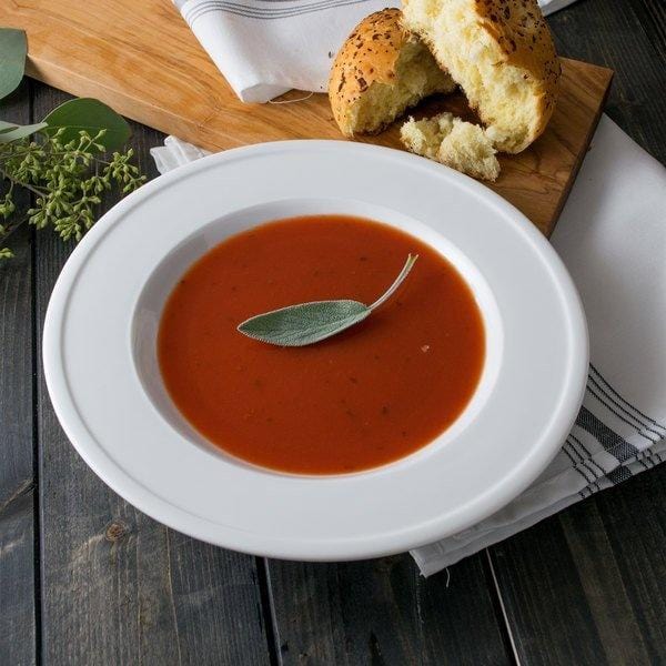 Un bol blanc de soupe à la tomate rouge sur une table en bois noir. A l’arrière du pains coupés sur une planche en bois