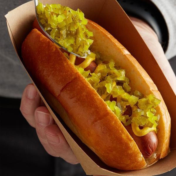 Un hot-dog dans un petit carton qui est tenu par une main, on y voit une sauce jaune en zig zag et une cuillère avec des petits carrés de cornichons 