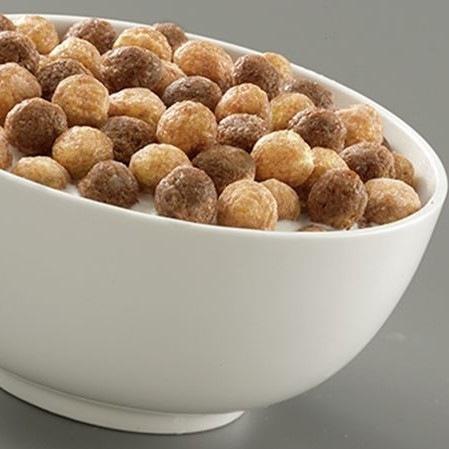 Un bol en verre blanc rempli de céréales en forme de boules brunes et beige avec du lait, le tout sur une table grise