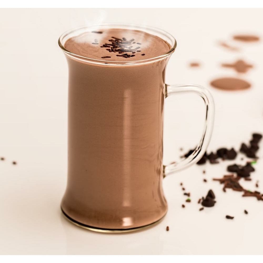 Une tasse transparente remplie de cacao chaud avec au-dessus des vermicelles de chocolats, le tout sur une table beige avec des copeaux de chocolat à droite