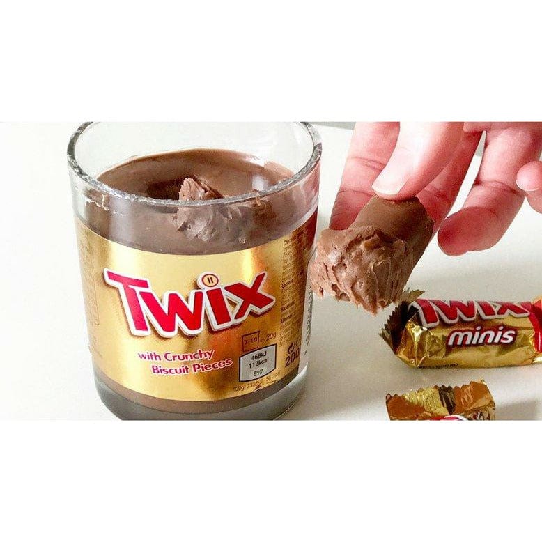Un pot en verre ouvert remplie d’une pâte brune avec une étiquette dorée, à droite il y a une main qui tient un Twix avec la pâte brune. Le tout sur une table blanche